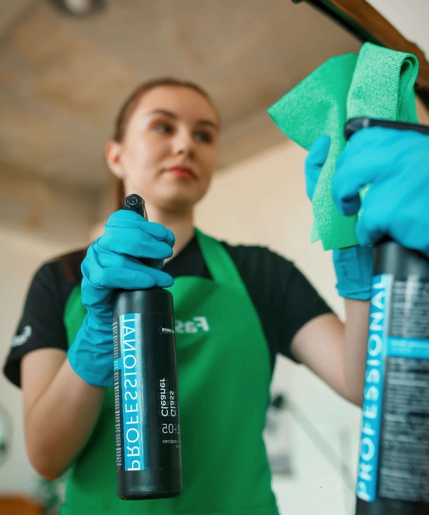 Сотрудница в синих перчатках протирает зеркало салфеткой и держит флакон с распылителем, флакон отражается в зеркале